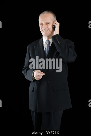 An agreeable businessman wearing a black suit smiling while speaking on mobile phone, on black background Stock Photo