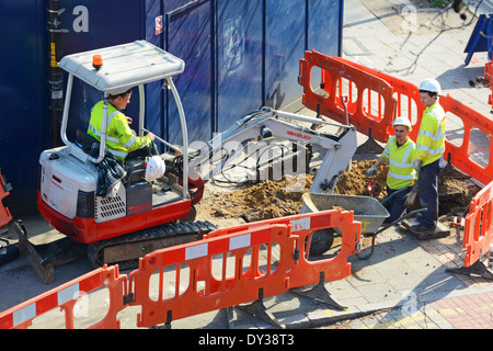 Aerial view three man team wearing hard hat & high visibility jacket using mini digger excavator digging up pavement behind red barriers London UK Stock Photo