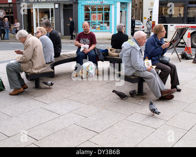 Feral pigeons eating scraps dropped by people sat on public bench, Truro, Cornwall, UK Stock Photo