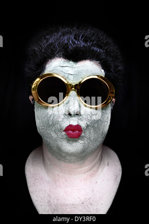 A woman wearing a green mud mask, red lipstick, and whimsical gold sunglasses puckers her lips Stock Photo
