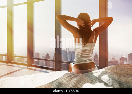 Young woman sitting on bed, stretching, overlooking city Stock Photo