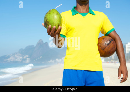 Brazilian soccer player in Brazil colors holding vintage brown football and drinking coconut Rio de Janeiro Stock Photo