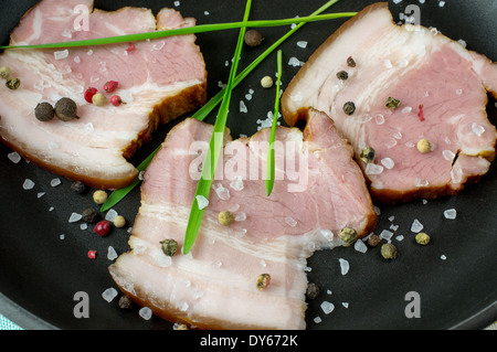 Pieces of bacon on a black frying pan with spices, salt and herbs. Stock Photo