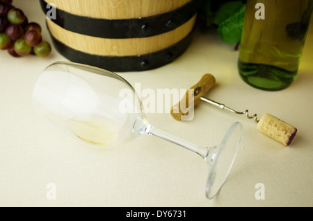 White wine in a glass lying. Background wine barrel, bottle of white wine, corkscrew, cork and grapes. Stock Photo
