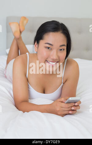 Smiling asian woman lying on bed texting on phone Stock Photo