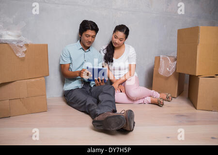 Happy couple sitting on floor using tablet surrounded by boxes Stock Photo