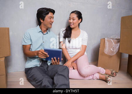 Happy couple sitting on floor using tablet surrounded by boxes Stock Photo