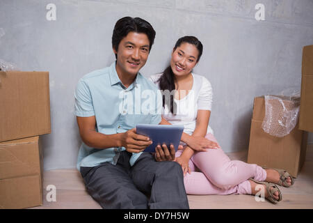 Happy couple sitting on floor using tablet surrounded by boxes Stock Photo