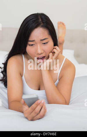 Shocked woman lying on bed holding smartphone Stock Photo