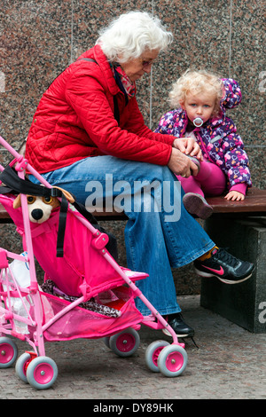 Grandmother and granddaughter senior woman and toddler young and old pink pram child pacifier girl Grandparent putting on a shoe take care of Stock Photo