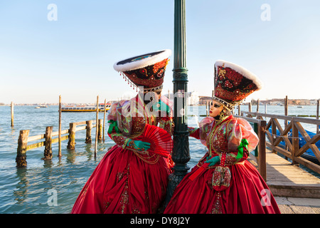 Venice carnival - two women in beautiful red fancy dress costumes, masks and hats pose by the lagoon, Carnevale di Venezia, Italy Stock Photo