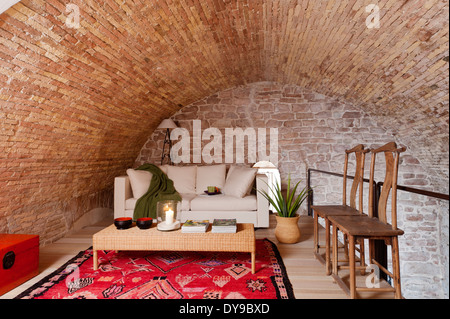 Vaulted stone walls in mezzanine space with white sofa bed, wicker coffee table and bright red ethnic rug Stock Photo