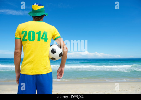 Brazilian soccer player in Brazil colors 2014 shirt and hat holding football tropical beach Stock Photo