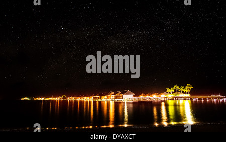 Night on tropical resort, many little beach houses glowing bright yellow lights in dark night, starry sky over sea Stock Photo