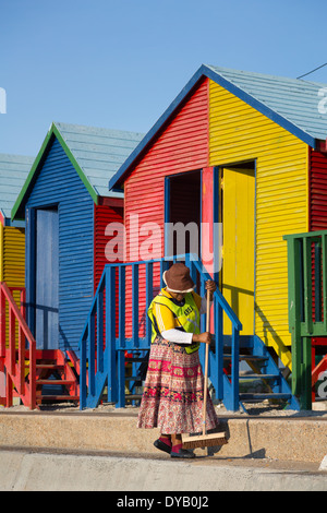 A street sweeper cleans the path in front of the iconic St James' beach huts. She is dressed in a colorful skirt Stock Photo
