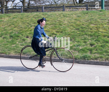 Panned shot of smiling woman dressed in period costume riding early bicycle Beamish Museum north east England UK Stock Photo