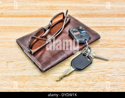A set of car keys and tinted sunglasses atop a brown leather wallet on wood tabletop. Stock Photo