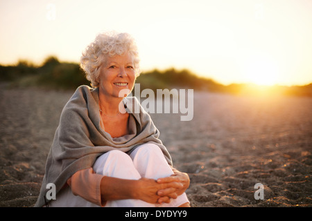 Portrait of smiling old woman sitting on the beach. Senior caucasian female relaxing outdoors Stock Photo