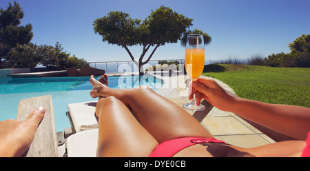 Young lady in bikini holding orange juice glass while sitting on a lounge chair along the poolside on a sunny day. Panorama view Stock Photo