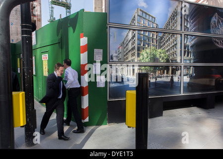 Two businessmen pass close to each other, next to a construction hoarding for a future office building. Stock Photo