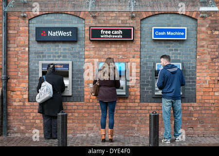 Three people at adjacent cash machines, one man watching the others. Stock Photo