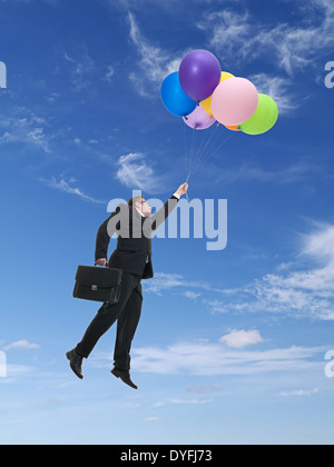 Businessman wearing black suit holding suitcase flying in the air being raised by bunch of colorful balloons Stock Photo