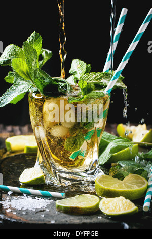 Glass of mojito cocktail with pouring rum and soda, fresh mint, limes and ice cubes over black background.