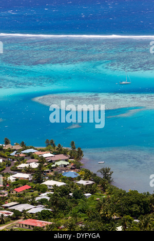 Sailboat anchored in a coral reef off the coast of Moorea with roofs of neighborhood houses on shore Stock Photo