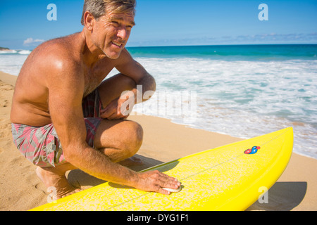 Middle-aged surfer waxing up his surfboard before heading into the surf, Hawaii Stock Photo