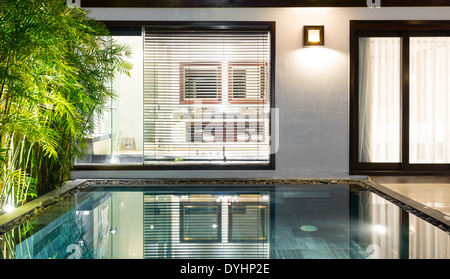 Hotel suite with pool inside. Door to bathroom in background. Windows with curtains and blinds. Palms near big swimming pool. Stock Photo