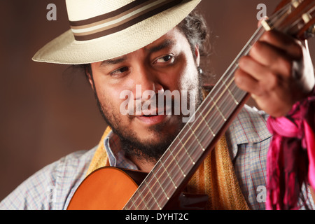 A close-up portrait of a man with a beard and dark curly hair wearing a light-colored straw fedora with brown and beige stripes. He is holding and playing an acoustic guitar, his left hand positioned Stock Photo