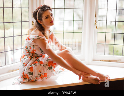 Young woman on a window sill in and old house, lit by natural light, while in a Fifties style summer dress. Stock Photo