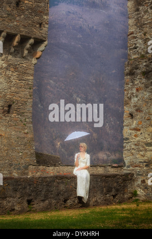 a woman in a white period dress is sitting on a stone wall with a parasol Stock Photo