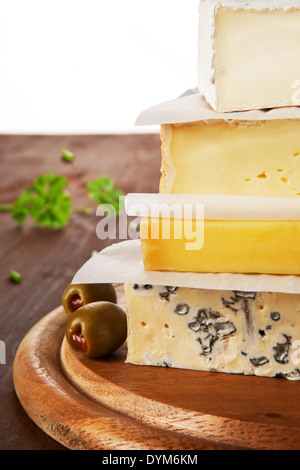 Various cheese pieces arranged on each other on brown wooden natural background with olives and parsley in background. Stock Photo