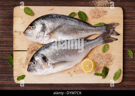 Two raw freash sea bream fish on wooden cutting board with cooking ingredients, fresh herbs basil, salt crystal and lemon. Stock Photo