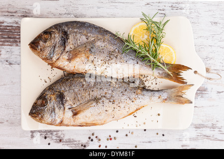 Delicious grilled sea bream fish on kitchen board with rosemary, lemon and colorful peppercorns on white textured background Stock Photo