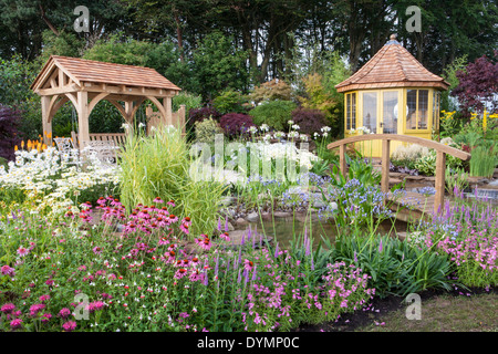 English country cottage garden arbour in summer with bridge summerhouse summer house cabin small pond water feature a mixed colourful flower border UK Stock Photo