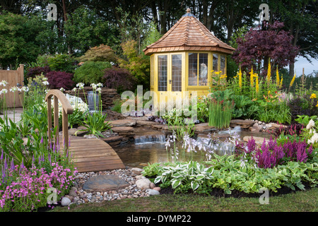 English country cottage garden summer with bridge summerhouse summer house cabin small pond water feature waterfall a mixed colourful flower border UK Stock Photo