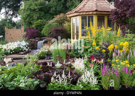 English country cottage garden summer with summerhouse summer house cabin small pond water feature waterfall mixed colourful flower border borders UK Stock Photo