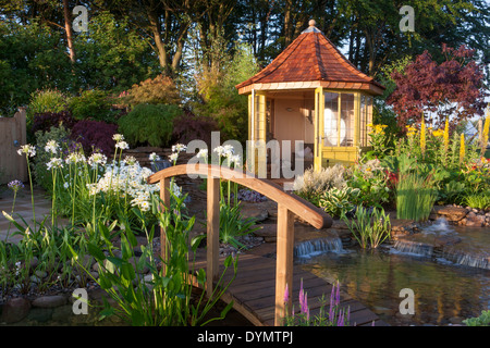 A country garden in summer with a bridge over to summerhouse summer house cabin small pond water feature waterfall a mixed colourful flower border UK Stock Photo
