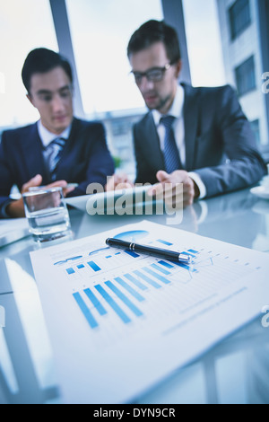 Image of business objects on background of two young businessmen discussing document in touchpad at meeting Stock Photo