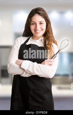 Female cooker with a black apron, smiling and holding a egg-beater, in a kitchen Stock Photo