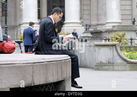 London, England, UK. Young Asian businessman reading newspaper in front of the Royal Exchange, Stock Photo