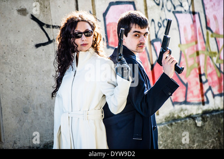 A couple of young fresh faced assassins holding guns pistols, killers, pair, man woman, team , standing back to back Stock Photo