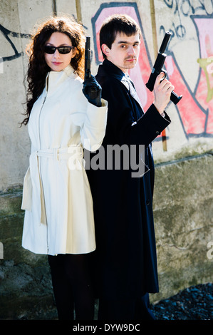 A couple of young fresh faced assassins holding guns pistols, killers, pair, man woman, team , standing back to back Stock Photo