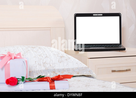 open laptop with a blank screen is on the nightstand in the bedroom and in front of him on the bed lay a flower and gift boxes Stock Photo