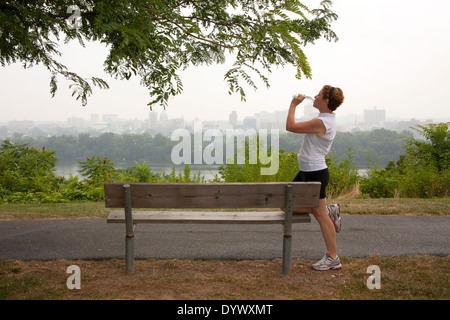 Mature woman taking a break and drinking water from jogging on a park bench overlooking the city Stock Photo