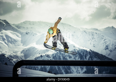 Snowboarder in woollen hat and camouflage trousers performs a tail grab aerial jump with dramatic mountains in the background. Stock Photo