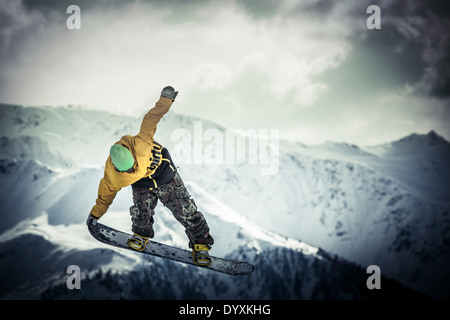 Snowboarder in woollen hat and camouflage trousers performs a tail grab aerial jump with dramatic mountains in the background. Stock Photo