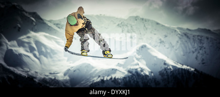 Snowboarder in woollen hat and camouflage trousers performs a tail grab aerial jump with dramatic mountains in the background. Stock Photo